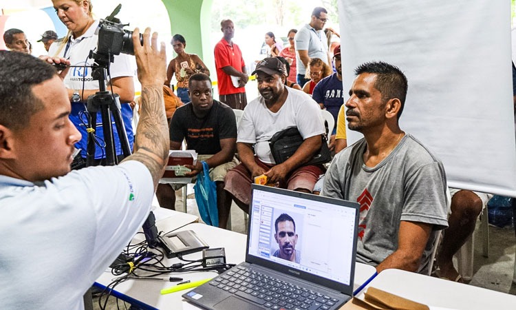 Funcionária do Detran tira foto de homem para carteira de identidade. Ao fundo outras pessoas aguardam atendimento