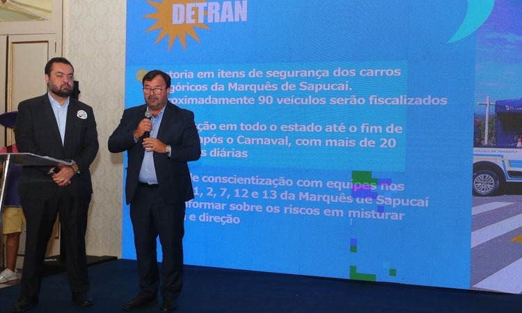 Governador do Rio de Janeiro observa fala de Vice-Presidente do Detran, ao microfone, com imagem de slide projetada ao fundo