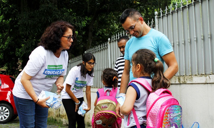 Duas funcionárias do Detran entregam blocos de colorir em calçada para duas crianças com mochilas rosa nas costas