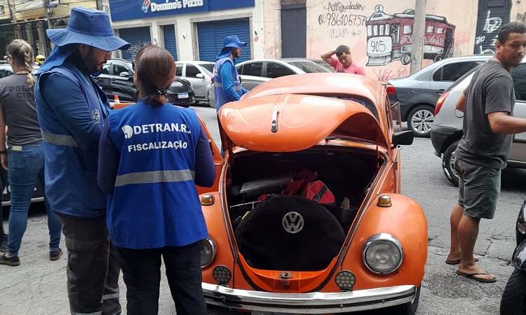 Dois agentes de fiscalização do Detran verificam fusca da cor laranja com capô aberto