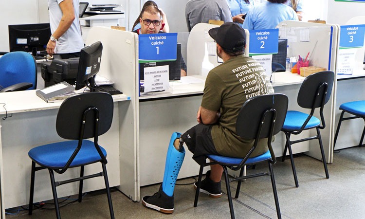 Funcionário do Detran atende um homem com perna mecânica, que aguarda sentado atrás do guichê