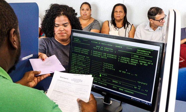 Funcionário do Detran atendendo mulher, que entrega formulário preenchido, em Posto do Detran 