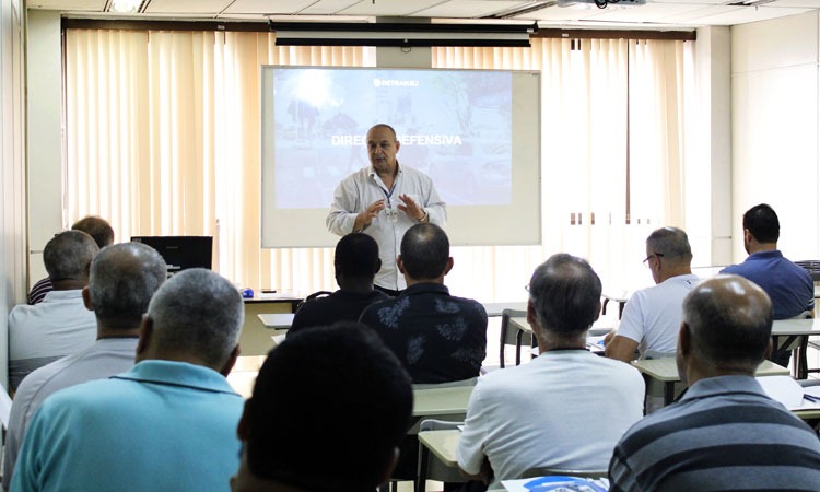 Instrutor do Detran dá aula para onze pessoas em sala de aula com projeção em tela ao fundo