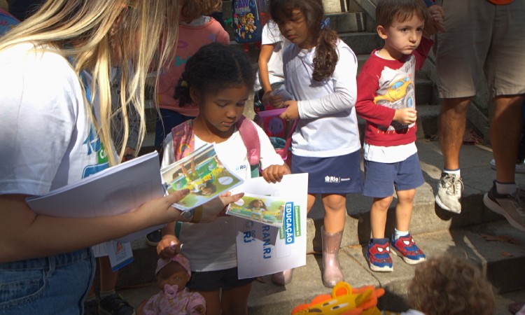 Crianças recebem bloquinho de colorir de funcionária do Detran,  em frente à escola