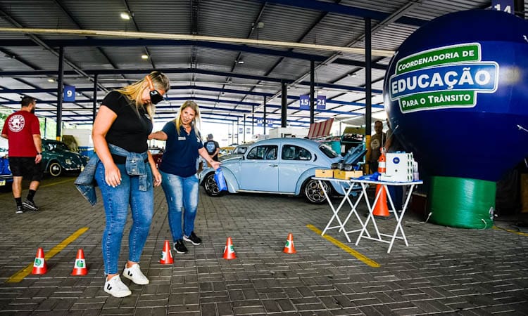 Mulher participa de atividade com óculos do Detran que simulam reações ao dirigir sob o efeito de com álcool, drogas e sono. Ao fundo alguns fuscas 
