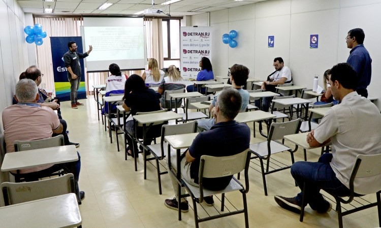 Instrutor do Detran dá aula para treze pessoas em sala de aula com projeção em tela ao fundo