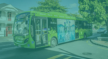 Ônibus verde com propaganda em sua lateral circula numa rua de pouco movimento.